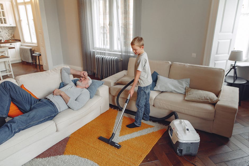 A young boy with light brown hair, wearing a light grey t-shirt, blue jeans, and black socks, stands upright while using a vacuum cleaner to clean a beige fabric sofa in a living room. An older man wi
