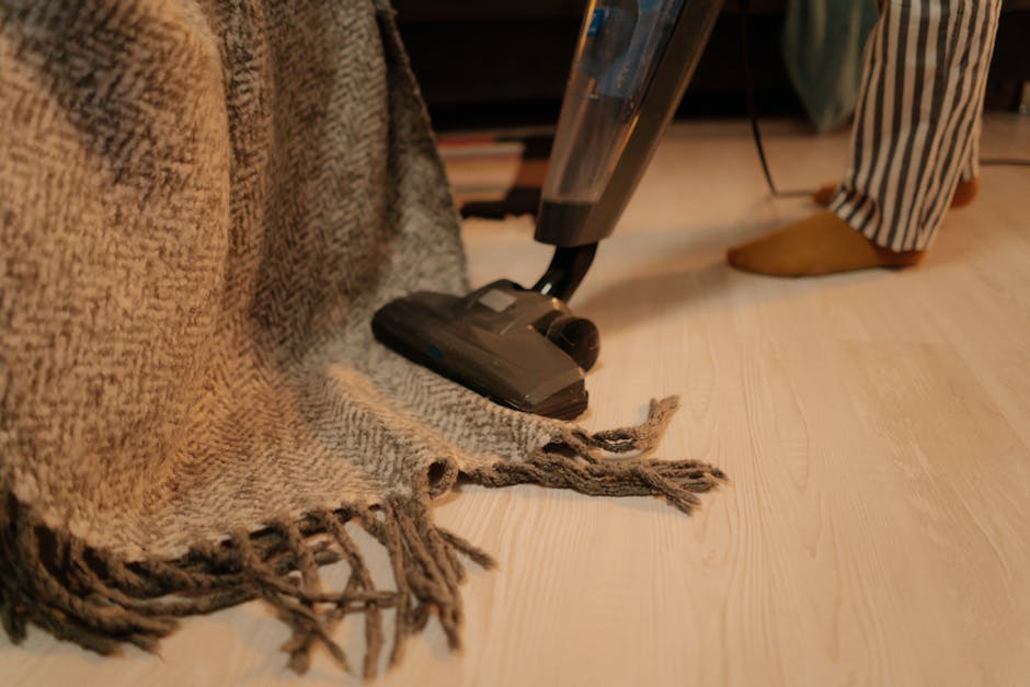 A young boy with light brown hair, wearing a light grey t-shirt, blue jeans, and black socks, stands upright while using a vacuum cleaner to clean a beige fabric sofa in a living room. An older man wi