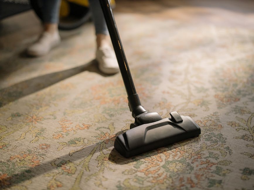 A person is vacuuming a decorative area rug with a black handheld vacuum cleaner, likely performing surface cleaning or deep cleaning to maintain hygiene and cleanliness in a residential or commercial space. The rug features intricate floral and leaf patterns in muted tones, laid out on a wooden floor, with natural light illuminating the scene. The vacuum cleaner's nozzle is pressed gently onto the rug's surface, and the person's feet in sneakers are visible in the background, indicating active cleaning. This image highlights the importance of regular sanitisation and maintenance of textiles as part of professional domestic cleaning services offered by Carpet Cleaners W4, based on the visible context of surface cleaning tips for locals around Chiswick High Road, W4.